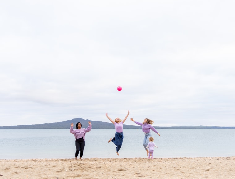 HELP Auckland spread the word page. 
Three women and a child smiling and playing with a ball on a beach.  In the background is the ocean and Rangitoto Island.