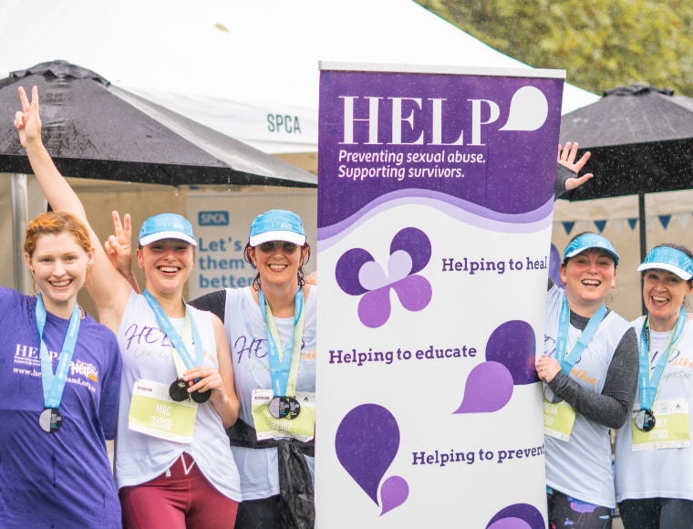 HELP Auckland Marathon home page.  Group of five smiling women next to HELP Auckland sign. They are wearing medals and have been running in the Auckland Marathon to raise money for Help Auckland.  