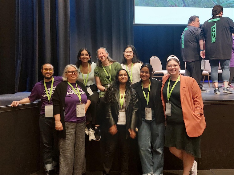 A group of committed Embassadors who look after the safe online community called Dear Em. They are standing together and smiling alongside HELP Auckland staff at a conference.